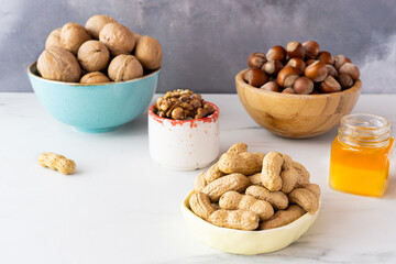 Fototapeta na wymiar Cashews in shell, hazelnuts in shell, walnuts in shell and peeled in different bowls are on a white table against a gray wall. There is a small jar of honey nearby. Natural organic products concept