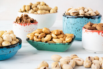 Peeled cashews in a green rectangular bowl stand on a white background. There are various nuts (hazelnuts, almonds, walnuts) in bowls nearby. Pistachios are scattered in the lower right corner