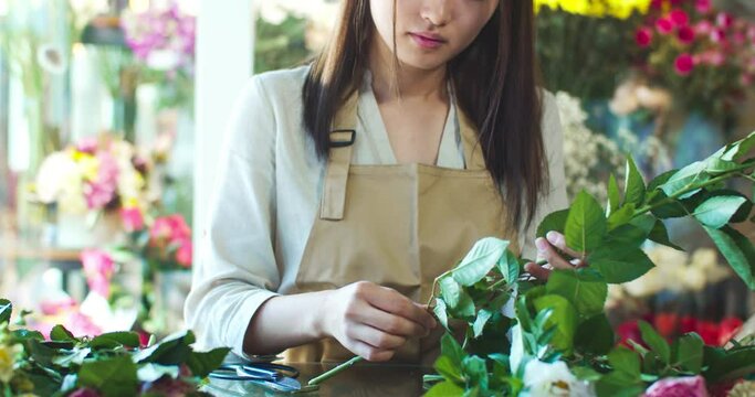 Close up of charming Asian woman florist working in flower store. Gentle hands of good-looking female worker taking blossoms and cutting stems of roses. Business, working process concept.