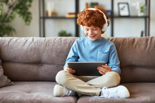Delighted redhead boy with tablet sitting on sofa.