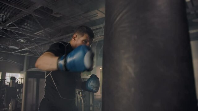 Young professional athlete strikes a punching bag aggressively with great force while wearing gloves. Smoky gym with lots of exercise equipment.