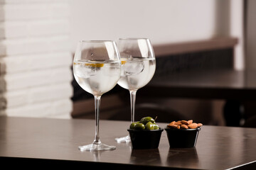 large glasses with alcoholic drink, ice and a piece of natural orange and container with olives and almonds, on top of the wooden table, inside the restaurant and free space for decoration.