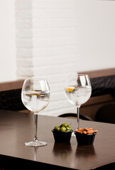 large glasses with alcoholic drink, ice and a piece of natural orange and container with olives and almonds, on top of the wooden table, inside the restaurant and free space for decoration.
