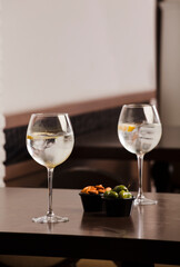 large glasses with alcoholic drink, ice and a piece of natural orange and container with olives and almonds, on top of the wooden table, inside the restaurant and free space for decoration.