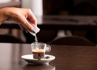 female hand with sugar envelope dumped into coffee cup, on top of wooden table, inside cafeteria and free space for decoration.