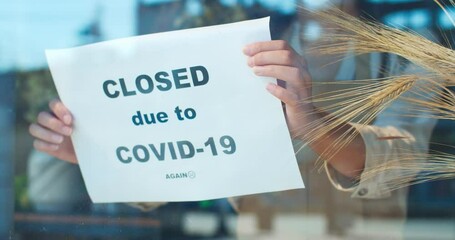 Front view of window of closing flower store during pandemic period. Young female florist in apron sticking sheet of paper with text "closed due to COVID-19". Quarantine, lockdown measures.