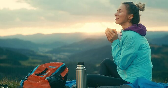 Woman hiker, hiking backpacker traveler camper in sleeping bag, fleabag drinking hot tea and relaxing on top of mountain. Health care, authenticity, sense of balance and calmness. Outdoor work. 4K