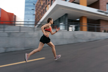 Obraz premium Young woman sprinting in the morning outdoors. Side view of female runner in motion working out in the city.