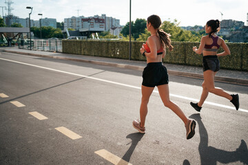 Fototapeta premium Young woman running on sidewalk in morning. Health conscious concept. Healthy active lifestyle. Active girls jogging together on road in the city.