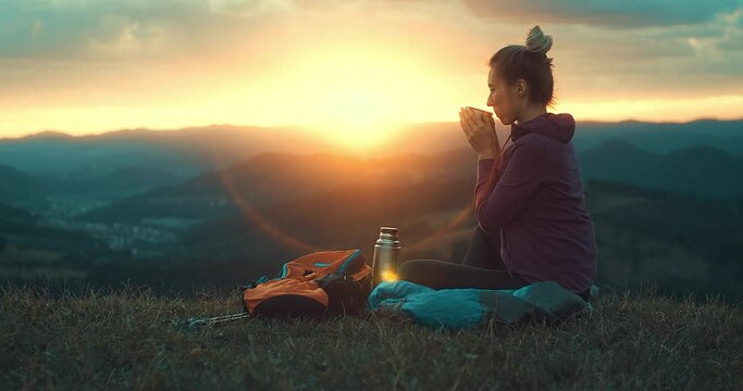 Woman hiker, hiking backpacker traveler camper in sleeping bag, fleabag drinking hot tea and relaxing on top of mountain. Health care, authenticity, sense of balance and calmness. Outdoor work. 4K