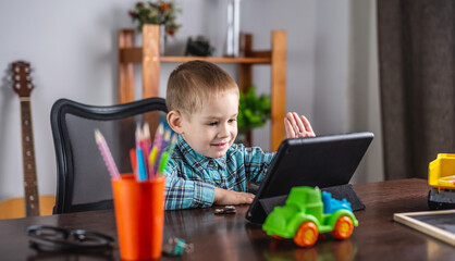 Small boy in a shirt is looking at the tablet screen and waving. Concept of online communication and distance education