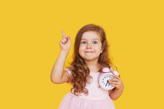 Time for a party. A pensive cute child in a fancy dress holds a small alarm clock in his hands and shows up. Yellow studio background..