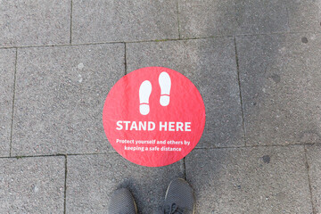Gray pair of shoes standing front of a red sign saying stand here to avoid spread of covid19....