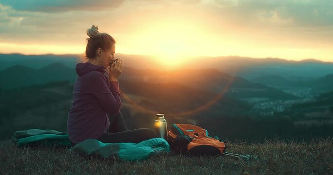 Woman hiker, hiking backpacker traveler camper in sleeping bag, fleabag drinking hot tea and relaxing on top of mountain. Health care, authenticity, sense of balance and calmness. Outdoor work. 4K