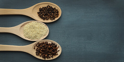 Three wooden spoons with spices lie on a dark wooden background. Selective focus. Banner. Various spices: cloves, black pepper and sesame.