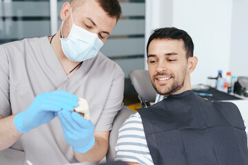 Male dentist in medical mask explaining artificial teeth to smiling patient in clinic