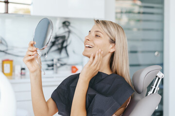 Young woman patient is looking in the mirror and admires her new smile after dental treatment in the dentistry clinic.