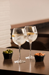 Two glasses of balloon, with ice and alcohol, pieces of orange and olives and almonds in a container, on the wooden table inside the restaurant and free space for decoration.