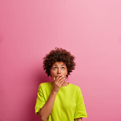 Vertical shot of young curly haired woman found out stunning news, has startled expression, covers...