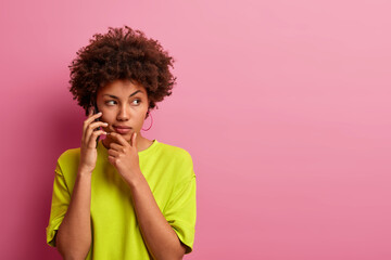 Photo of serious dark skinned woman holds chin, has telephone conversation, dressed in casual...