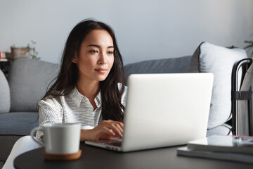 Young ambitious asian girl working remote from home, looking at laptop screen and smiling. Woman checking mail or researching while telecommuting, sitting on floor at her apartment
