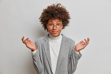 Elegant puzzled female boss wears formal clothes, spreads palms sideways, faces dilemma, shrugs shoulders, being unaware, poses against studio background. Human perceprion, hesitation concept