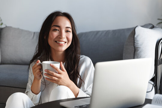 Home, Lifestyle And Leisure Concept. Beautiful Asian Girl Smiling And Drinking Tea While Watching Online Movies On Laptop, Enjoying Streaming Videos, Checking Out New Tv Series And Resting