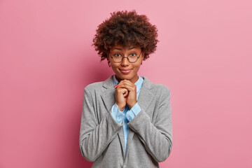 Isolated shot of dark skinned pretty woman in eyeglasses with curly hairstyle keeps hands under chin, listens attentively interlocutor, prepares for formal meeting, isolated on pink background.