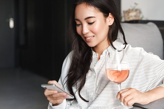Close-up of elegant young asian woman relaxing at home, browsing online shopping sites on mobile phone and enjoying glass of wine. Female resting on weekend, scrolling social networks