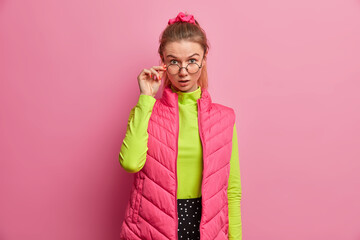 Portrait of shocked adult European woman looks through optical glasses, has astonished expression, keeps hand on rim of spectacles, surprised by what she saw, wears bright clothing, poses indoor