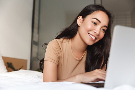Portrait of beautiful happy asian woman lying in bed relaxed and looking at laptop screen. Girl smiling happy while browsing internet on computer, resting at home