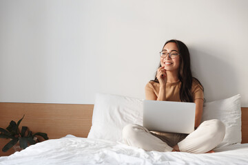 Work from home, freelance and lifestyle concept. Portrait of creative young asian female sitting on bed with laptop, smiling thoughtful and looking away while think-up. Girl shopping online - Powered by Adobe
