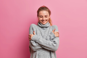 Obraz na płótnie Canvas Happy tender girl dressed in grey warm oversized sweater, embraces herself, enjoys comfort during rainy autumn day, wears transparent glasses smiles pleasantly, isolated over pink background