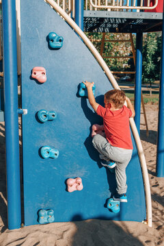 Little Preschool Boy In Red T-shirt Climbing Rock Wall At Playground Outside On Summer Day. Happy Childhood Lifestyle Concept. Seasonal Outdoors Activity For Kids. View From Back.