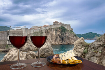 Two glasses of wine with cheese and meat assortment on view of the fortress walls, rocky coast and blue sea in Dubrovnik, Croatia. Stunning view of Dubrovnik city walls and Dubrovnik old town.