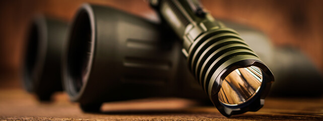 Banner detail close up of army - hunting binocular and flashlight on a blurred wooden background. 
