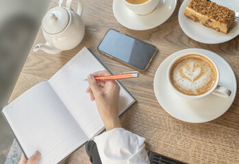 The girl writes notes in her diary with an orange pen over a Cup of tea.