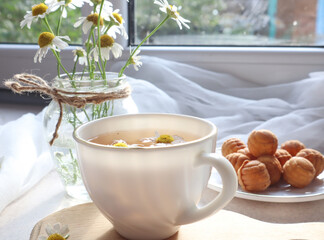  A Cup of fragrant chamomile tea with a delicate bouquet of daisies close-up on the background of the window-the concept of a pleasant tea party and pastime.