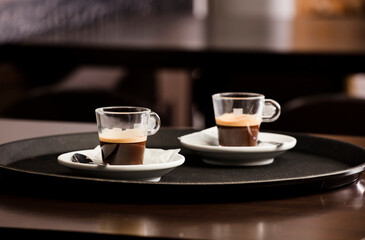 Dos tazas de café caliente recién hecho, en la bandeja del camarero, en la mesa de madera, dentro de la cafetería y espacio libre para decoración.