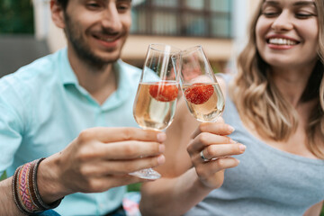 Happy couple portrait clinking two glasses with sparkling wine and strawberries inside with blurred house on background. Celebrating love