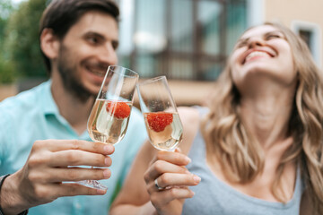 Happy couple portrait clinking two glasses with sparkling wine and strawberries inside with blurred house on background. Celebrating love