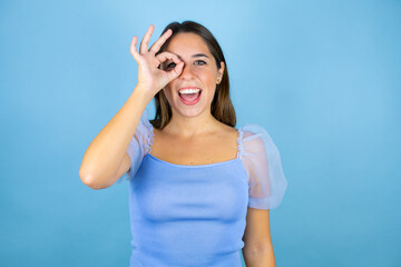 Young beautiful woman over isolated blue background doing ok gesture shocked with smiling face, eye looking through fingers