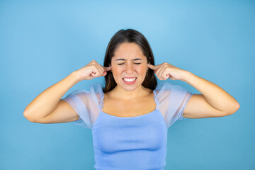 Young beautiful woman over isolated blue background covering ears with fingers with annoyed expression for the noise of loud music. Deaf concept.