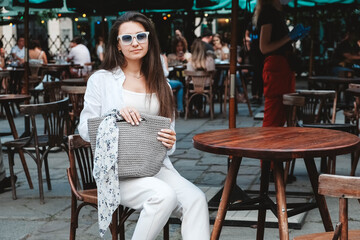 Beautiful young brunette woman wearing in sunglasses and white clothes, holding knitted bag sitting at the table of a street cafe. Copy, empty space for text