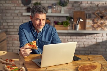Man working from home on laptop computer, sitting at table in kitchen, eating online ordered pizza.