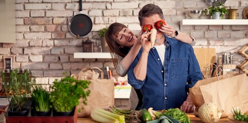 Happy couple in the kitchen unpacking grocery bags full of vegetables arriving home from shopping having fun, screw around.