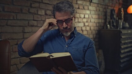 Mature man in his 50s reading book at home sitting in dark living room.  