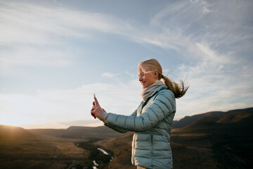 Young caucasian blonde female taking selfie on top of mountain while hiking at sunset.