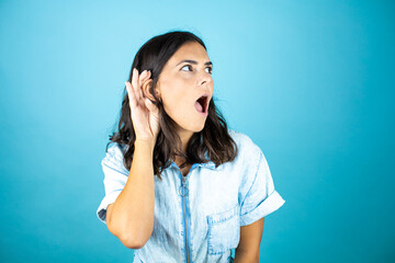 Young beautiful woman wearing a denim jumpsuit over isolated blue background surprised with hand over ear listening an hearing to rumor or gossip.