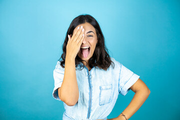 Young beautiful woman over isolated blue background covering one eye with hand, confident smile on face and surprise emotion.